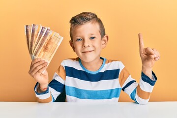 Adorable caucasian kid holding 500 norwegian krone banknotes sitting on the table smiling with an idea or question pointing finger with happy face, number one