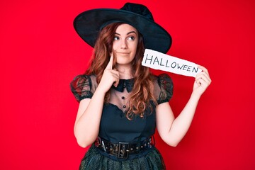 Young beautiful woman wearing witch costume holding paper with halloween message serious face thinking about question with hand on chin, thoughtful about confusing idea