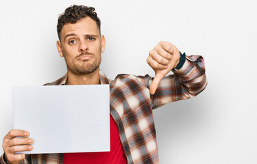 Young hispanic man holding blank empty banner with angry face, negative sign showing dislike with...