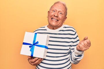 Senior handsome grey-haired man holding birthday gift over isolated yellow background screaming proud, celebrating victory and success very excited with raised arm