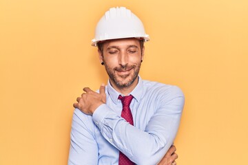 Young handsome man wearing architect hardhat hugging oneself happy and positive, smiling confident. self love and self care