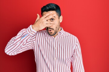 Young hispanic man wearing business shirt peeking in shock covering face and eyes with hand,...
