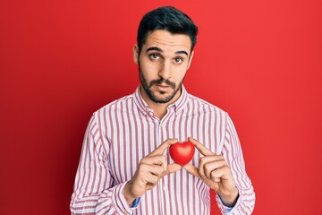 Young hispanic man holding heart clueless and confused expression. doubt concept.