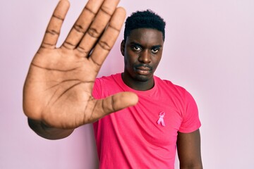 Handsome black man wearing pink cancer ribbon attached on shirt with open hand doing stop sign with...