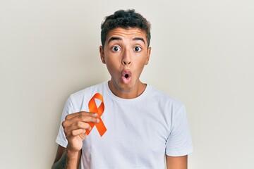 Young handsome african american man holding awareness orange ribbon scared and amazed with open...