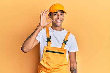 Young handsome african american man wearing handyman uniform over yellow background smiling positive doing ok sign with hand and fingers. successful expression.