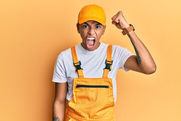 Young handsome african american man wearing handyman uniform over yellow background angry and mad raising fist frustrated and furious while shouting with anger. rage and aggressive concept.