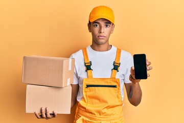 Young handsome african american man holding delivery package and showing smartphone screen relaxed with serious expression on face. simple and natural looking at the camera.