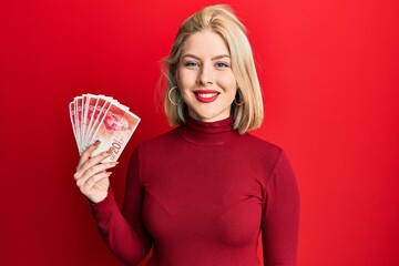 Young blonde woman holding 20 israel shekels banknotes looking positive and happy standing and smiling with a confident smile showing teeth