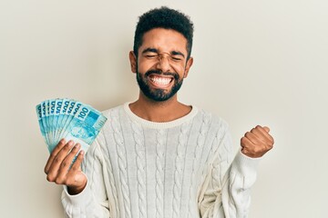 Handsome hispanic man with beard holding 100 brazilian real banknotes screaming proud, celebrating...