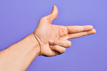 Hand of caucasian young man showing fingers over isolated purple background gesturing fire gun weapon with fingers, aiming shoot symbol