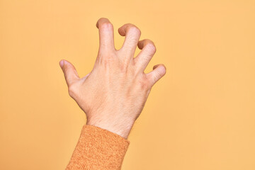 Hand of caucasian young man showing fingers over isolated yellow background grasping aggressive and scary with fingers, violence and frustration