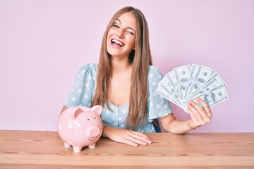 Young blonde girl holding dollars and piggy bank sitting on the table winking looking at the camera...