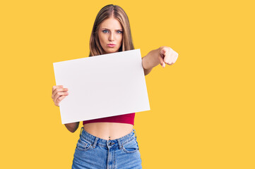Fototapeta na wymiar Young beautiful blonde woman holding blank empty banner pointing with finger to the camera and to you, confident gesture looking serious