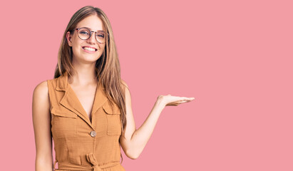 Young beautiful blonde woman wearing casual clothes and glasses smiling cheerful presenting and pointing with palm of hand looking at the camera.