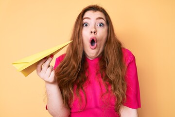 Young redhead woman holding paper airplane scared and amazed with open mouth for surprise,...