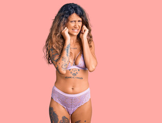 Young hispanic woman with tattoo wearing lingerie covering ears with fingers with annoyed expression for the noise of loud music. deaf concept.