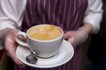 The waiter's gloved hands hold a cup of freshly brewed coffee with foam. Cup close-up. Selective focus.