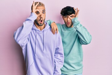 Homosexual gay couple standing together wearing casual clothes doing ok gesture shocked with surprised face, eye looking through fingers. unbelieving expression.