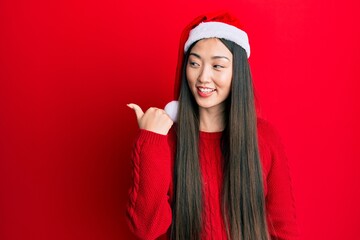 Young chinese woman wearing christmas hat pointing thumb up to the side smiling happy with open mouth