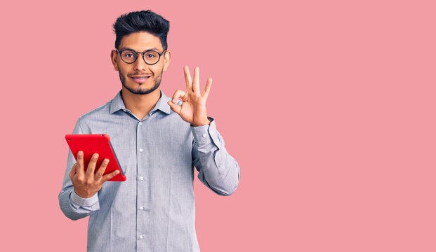 Handsome latin american young man holding touchpad doing ok sign with fingers, smiling friendly gesturing excellent symbol