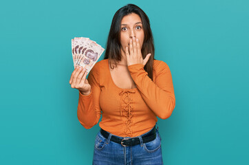 Beautiful hispanic woman holding mexican pesos covering mouth with hand, shocked and afraid for mistake. surprised expression