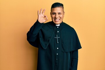 Young latin priest man standing over yellow background smiling positive doing ok sign with hand and...