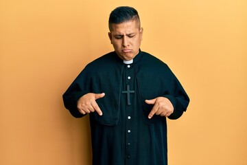 Young latin priest man standing over yellow background pointing down looking sad and upset,...