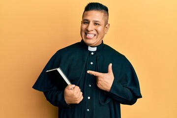 Young latin priest man holding bible smiling happy pointing with hand and finger