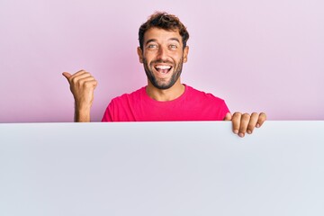 Handsome man with beard holding blank empty banner pointing thumb up to the side smiling happy with open mouth
