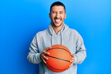 Handsome man with tattoos holding basketball ball sticking tongue out happy with funny expression.