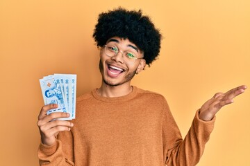 Young african american man with afro hair holding 50 thai baht banknotes celebrating achievement with happy smile and winner expression with raised hand