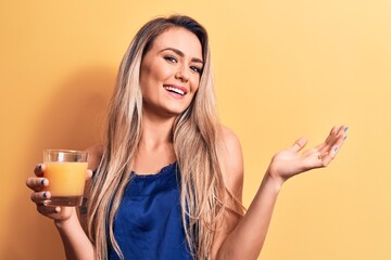 Young beautiful blonde woman drinking glass of healthy orange juice over yellow background celebrating achievement with happy smile and winner expression with raised hand