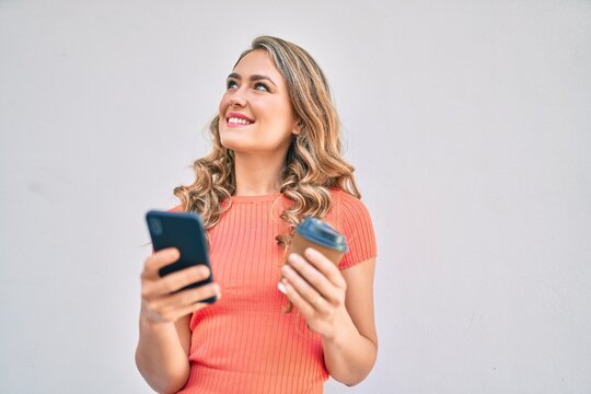 Young blonde girl smiling happy using smartphone and drinking take away coffee at the city.