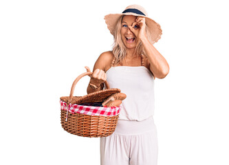 Middle age fit blonde woman wearing summer hat and holding picnic wicker basket with bread smiling happy doing ok sign with hand on eye looking through fingers