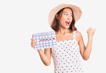 Obraz na płótnie Canvas Young beautiful chinese girl wearing summer hat holding heart calendar pointing thumb up to the side smiling happy with open mouth