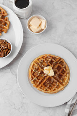 Homemade Pecan Waffles on a White Plate with a Small Dish of Pecans; One Pecan Waffle on a White Plate with Butter and Maple Syrup; Butter and Maple Syrup in Background