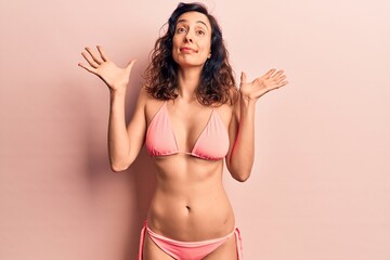 Young beautiful hispanic woman wearing bikini clueless and confused with open arms, no idea and...