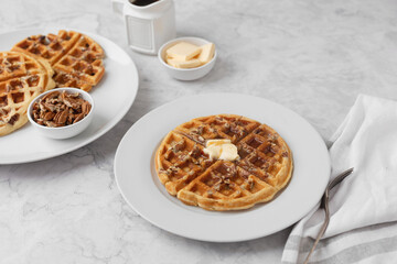 Homemade Pecan Waffles on a White Plate with a Small Dish of Pecans; One Pecan Waffle on a White Plate with Butter and Maple Syrup; Butter and Maple Syrup in Background