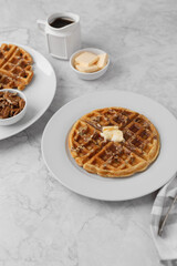 Homemade Pecan Waffles on a White Plate with a Small Dish of Pecans; One Pecan Waffle on a White Plate with Butter and Maple Syrup; Butter and Maple Syrup in Background