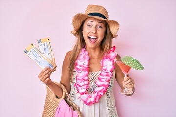 Middle age hispanic woman wearing hawaiian lei holding boarding pass and cocktail smiling and laughing hard out loud because funny crazy joke.