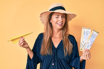 Middle age hispanic woman wearing summer hat holding boarding pass and paper airplane winking...