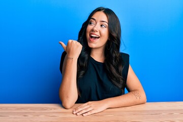 Beautiful brunette young woman wearing casual clothes sitting on the table pointing thumb up to the...