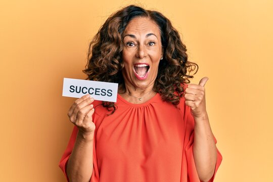 Middle age hispanic woman holding success message paper pointing thumb up to the side smiling happy with open mouth