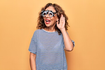 Middle age beautiful woman controlling vision using optometry glasses over yellow background...