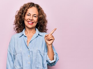 Middle age beautiful woman wearing casual denim shirt standing over pink background with a big smile on face, pointing with hand finger to the side looking at the camera.