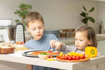 Cute little girl 2-4 in gray dress and boy 7-10 in T-shirt cooking pizza together in kitchen. Brother and sister cooking