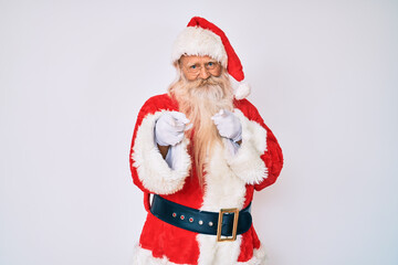 Old senior man with grey hair and long beard wearing santa claus costume with suspenders pointing fingers to camera with happy and funny face. good energy and vibes.