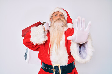 Old senior man with grey hair and long beard wearing santa claus costume holding shopping bags with open hand doing stop sign with serious and confident expression, defense gesture