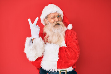 Old senior man with grey hair and long beard wearing traditional santa claus costume smiling with happy face winking at the camera doing victory sign. number two.